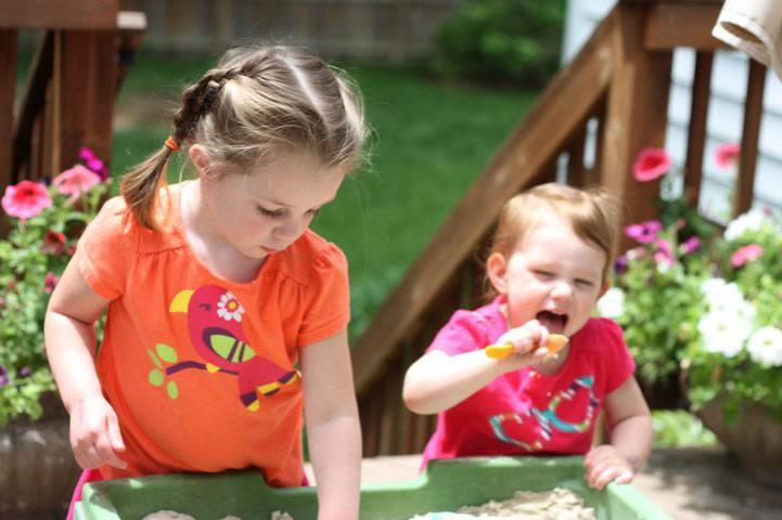 Two children are playing in a sandbox on a sunny day. One child is holding a small plastic shovel and attempting to scoop sand toward their mouth, while the other child digs in the sand.