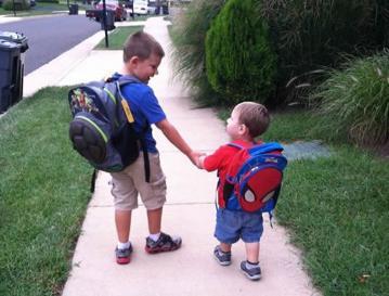 Two children wearing backpacks walk hand-in-hand down a sidewalk. The older child looks down at the younger child.